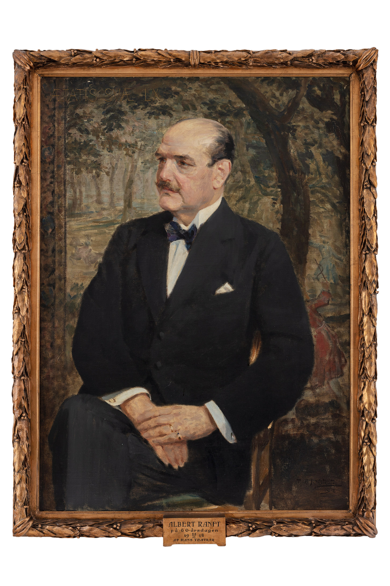 Albert Ranft sitter ner vänd mot vänster. Han är klädd i svart kostym med vit skjorta och svart fluga. Han har svart hår på sida an hjässan och mustasch. I bakgrunden en landskapsmålning.
