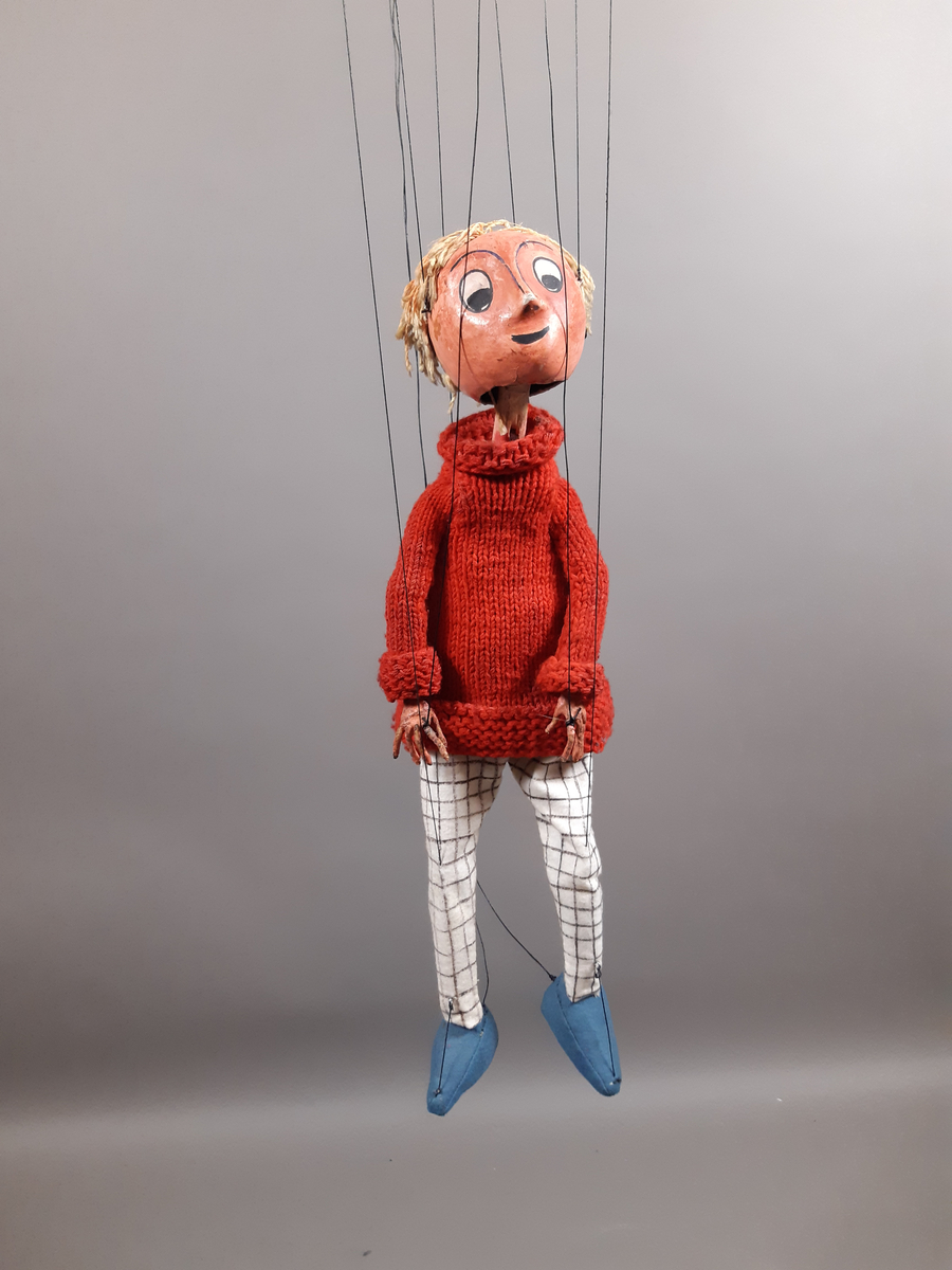 Marionettdocka föreställande "Kirre" en pojke med ljust hår, röd stickad polotröja, svartvitrutiga långbycor och blå skor.
