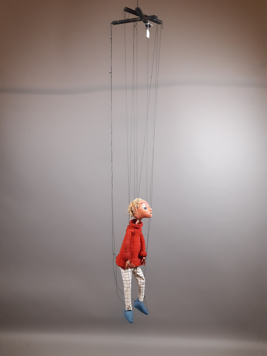 Marionettdocka föreställande "Kirre" en pojke med ljust hår, röd stickad polotröja, svartvitrutiga långbycor och blå skor.
