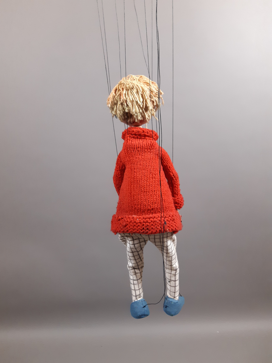 Marionettdocka föreställande "Kirre" en pojke med ljust hår, röd stickad polotröja, svartvitrutiga långbycor och blå skor.
