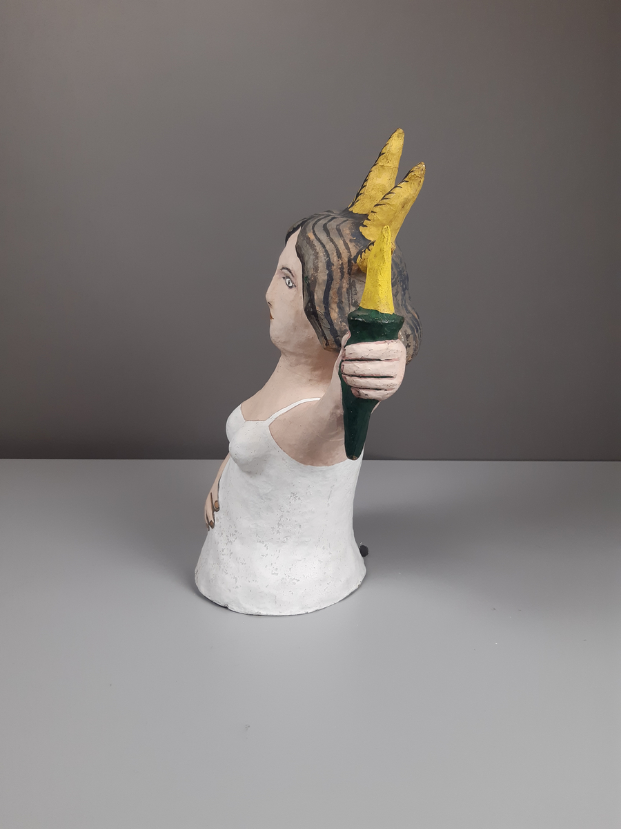 Docka i målad papier-maché, föreställande en kort knubbig kvinna med mörkt lockigt axellångt hår, hon är klädd i vit dräkt och har en gul krona och hållen en fackla i sin hand.
