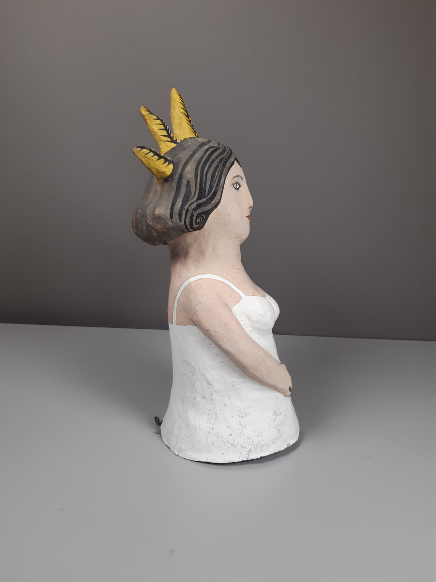 Docka i målad papier-maché, föreställande en kort knubbig kvinna med mörkt lockigt axellångt hår, hon är klädd i vit dräkt och har en gul krona och hållen en fackla i sin hand.
