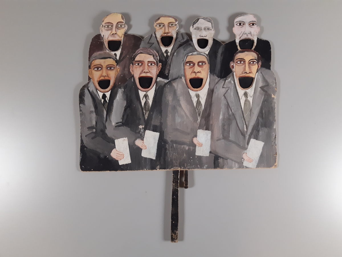 Åtta "pratande" män klädda i mörkgrå kostym målade på tjock kartong. Huvuden, hals och axlar på de fyra översta männen är utklippta. Männens munnar går att öppna och stänga med en träkonstruktion med två handtag på baksidan.
