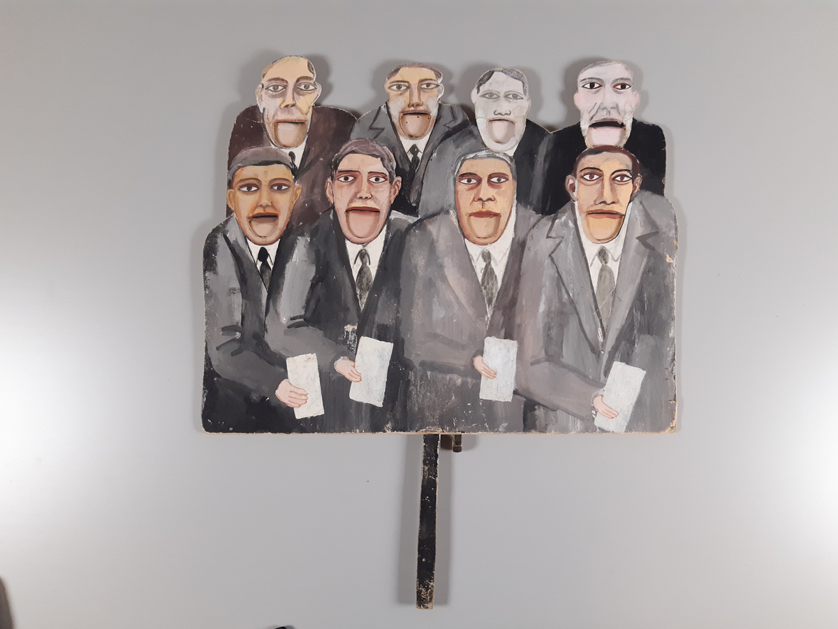 Åtta "pratande" män klädda i mörkgrå kostym målade på tjock kartong. Huvuden, hals och axlar på de fyra översta männen är utklippta. Männens munnar går att öppna och stänga med en träkonstruktion med två handtag på baksidan.
