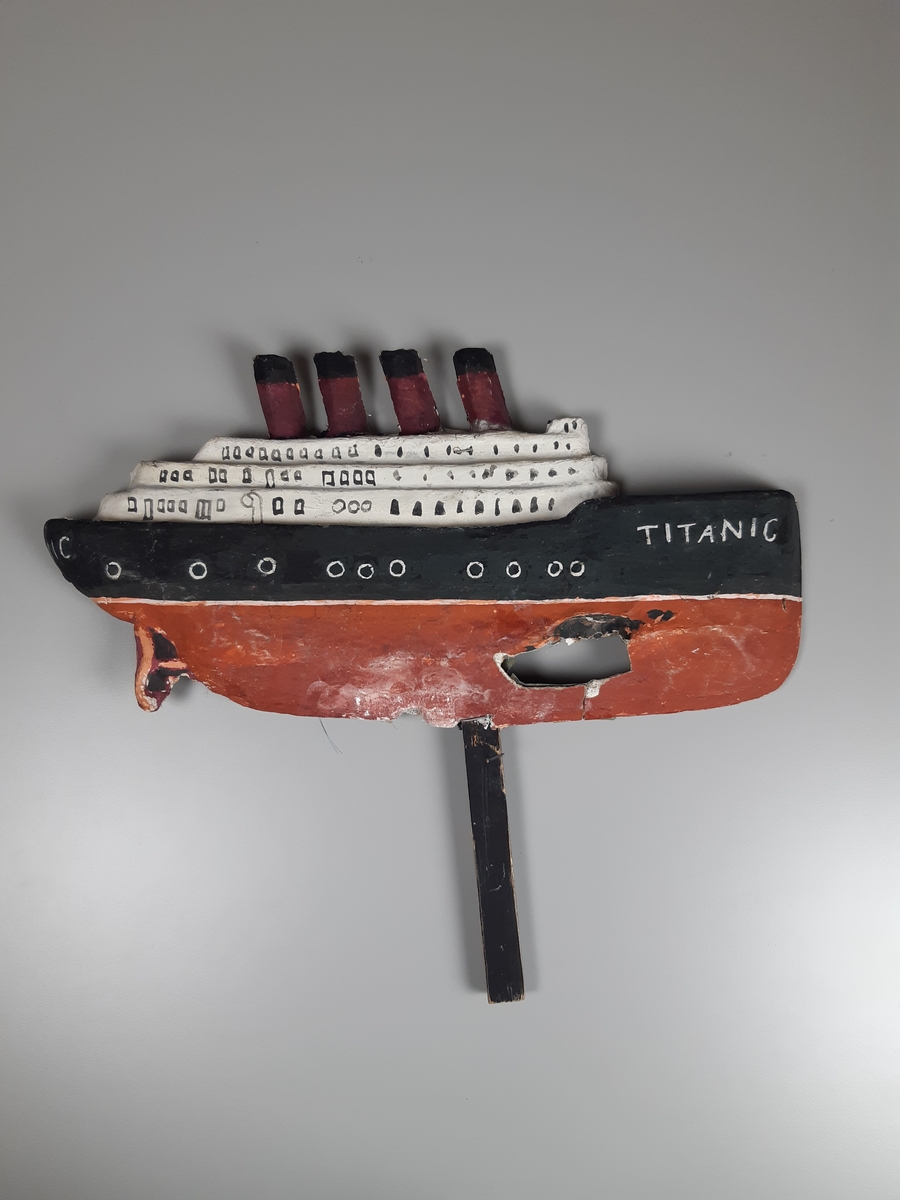 Relief i gips och papier-maché på ett skaft föreställande fartyget TITANIC i rött, svart och vitt. Fartyget har fyta höga skorstenar och ett hål i skrovet.
