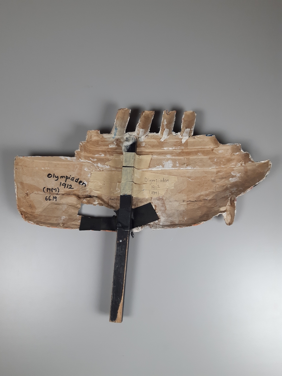 Relief i gips och papier-maché på ett skaft föreställande fartyget TITANIC i rött, svart och vitt. Fartyget har fyta höga skorstenar och ett hål i skrovet.
