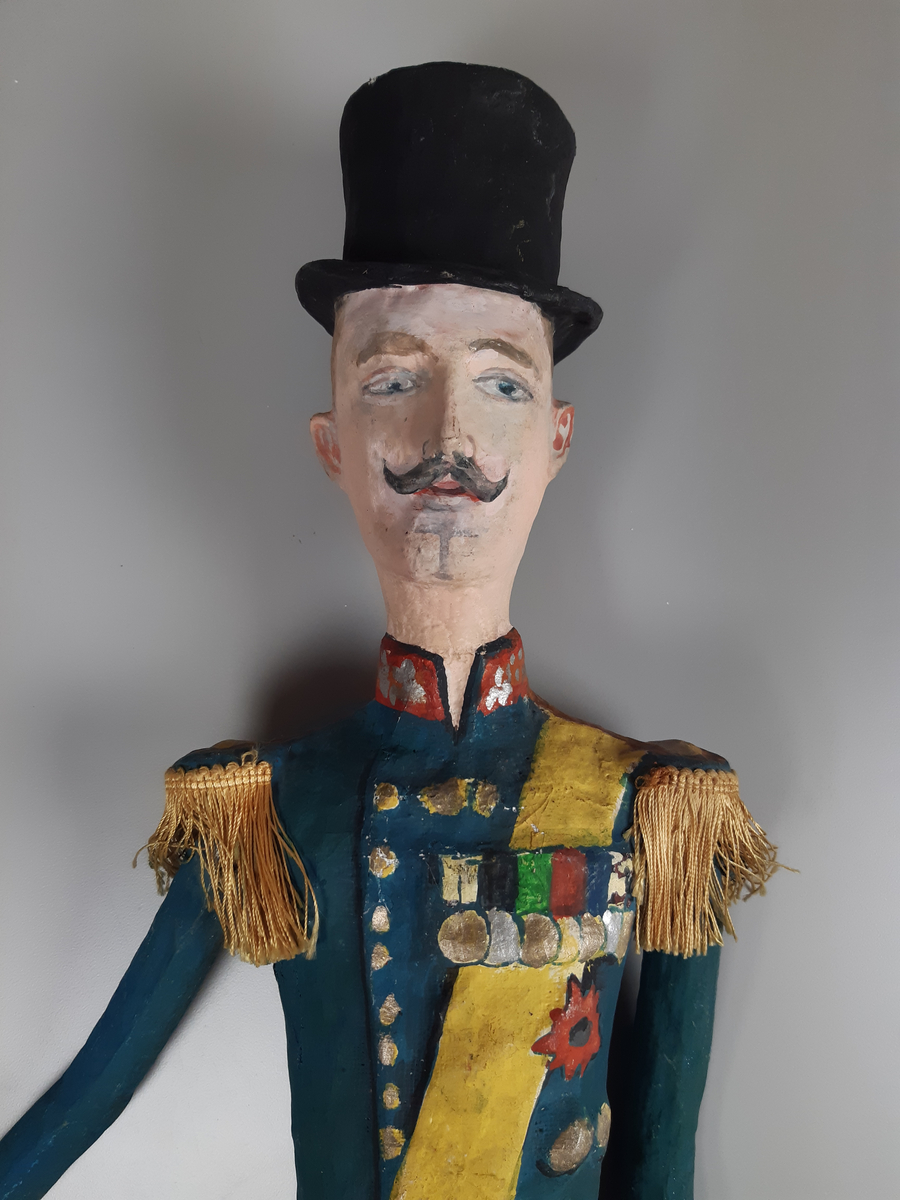 Docka i målad papier-maché, föreställande en äldre smal man i  hög svart hatt, grönblå uniform med röd ståkrage, gult ordensband och medaljer på bröstet, guld epoletter av snörfrans. Dockan har lösa armar och byxorna är i svart tyg.
