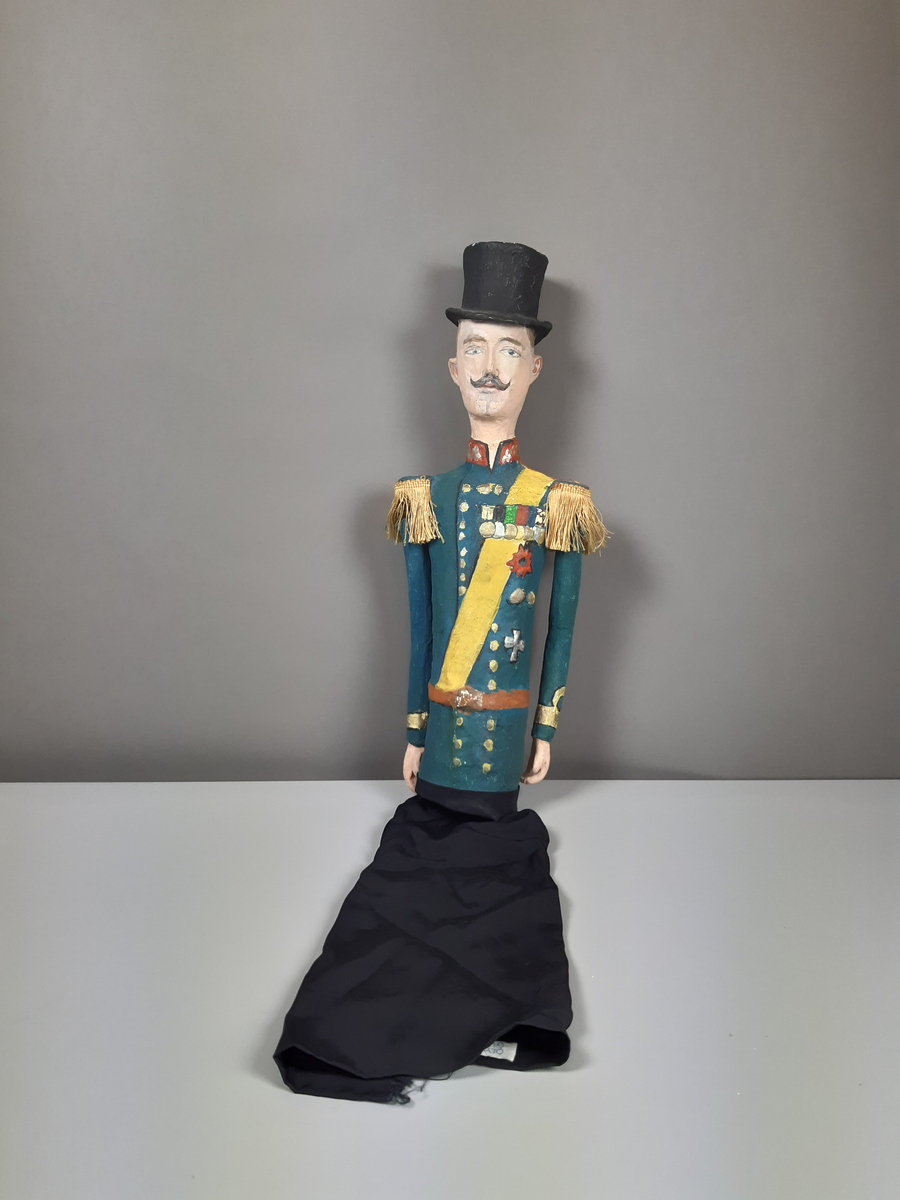 Docka i målad papier-maché, föreställande en äldre smal man i  hög svart hatt, grönblå uniform med röd ståkrage, gult ordensband och medaljer på bröstet, guld epoletter av snörfrans. Dockan har lösa armar och byxorna är i svart tyg.
