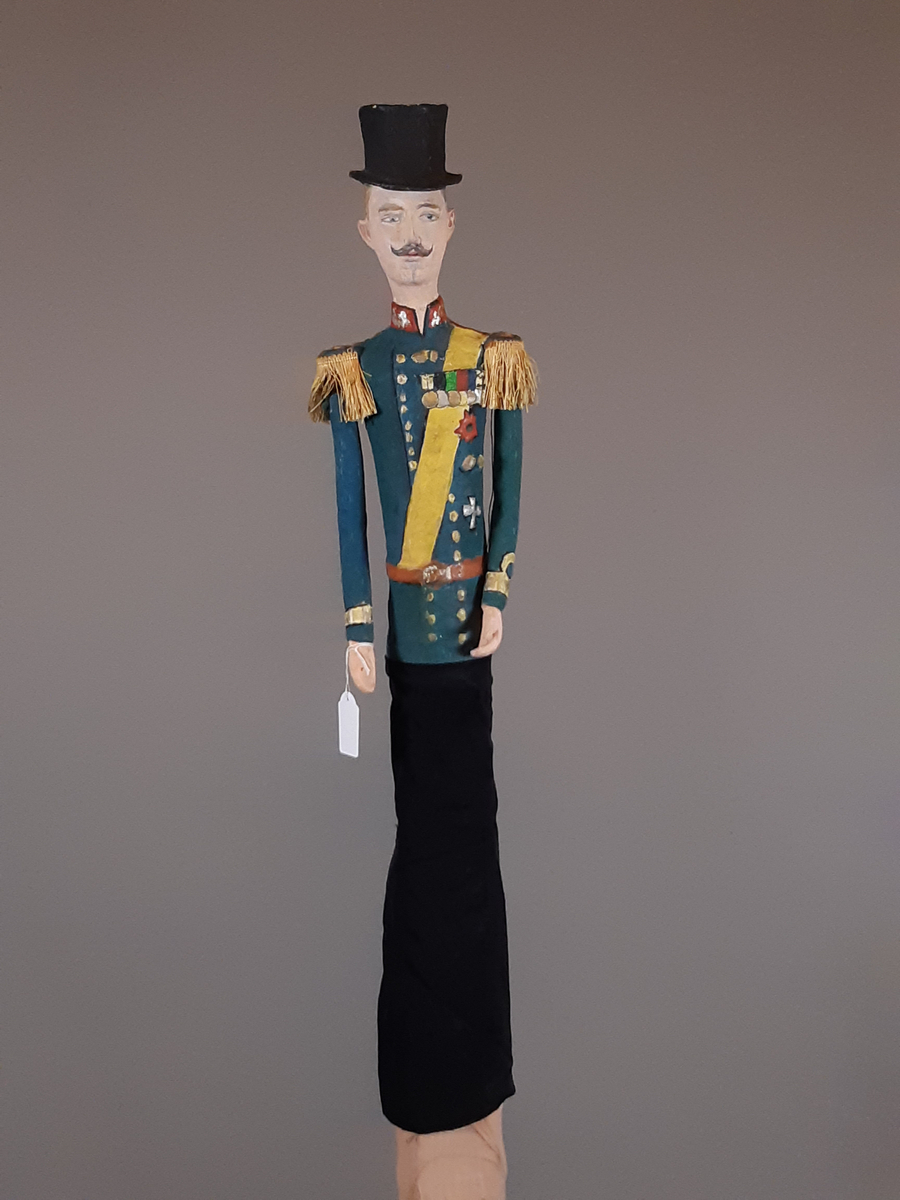 Docka i målad papier-maché, föreställande en äldre smal man i  hög svart hatt, grönblå uniform med röd ståkrage, gult ordensband och medaljer på bröstet, guld epoletter av snörfrans. Dockan har lösa armar och byxorna är i svart tyg.
