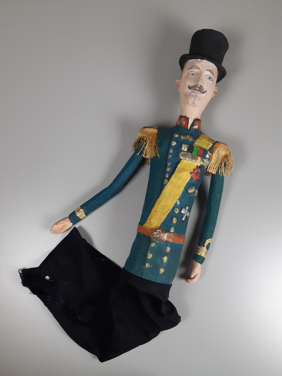 Docka i målad papier-maché, föreställande en äldre smal man i  hög svart hatt, grönblå uniform med röd ståkrage, gult ordensband och medaljer på bröstet, guld epoletter av snörfrans. Dockan har lösa armar och byxorna är i svart tyg.
