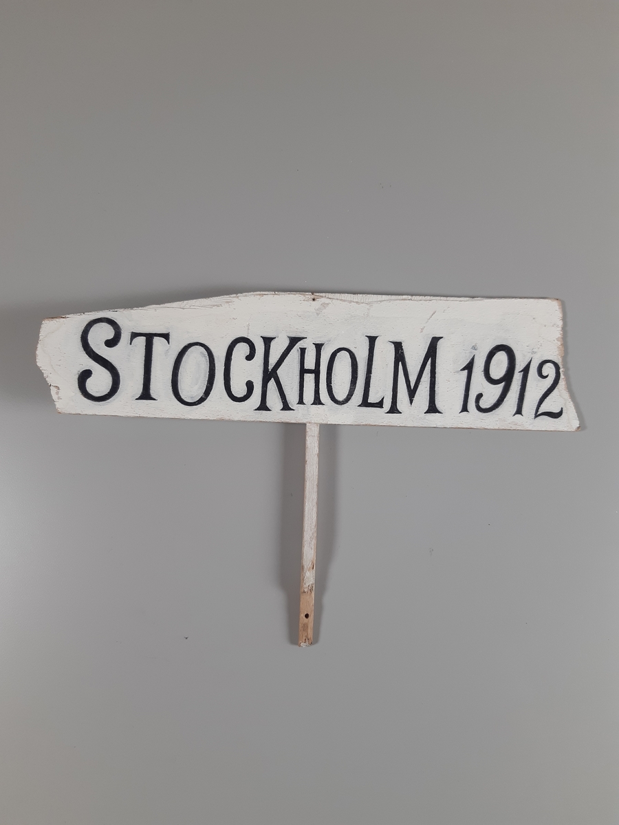 Vitmålad skylt med svarta bokstäver med skaft.
