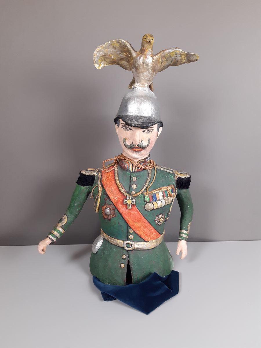 Docka i målad papier-maché, föreställande en man klädd i grön paraduniform och blank örnprydd hjälm. På bröstet har han en rad medaljer, ett rött ordensband över axeln, epåletter i guld med svarta tygfransar. Mannen har grått hår och mustasch. Byxorna består av mörkblå sammet.
