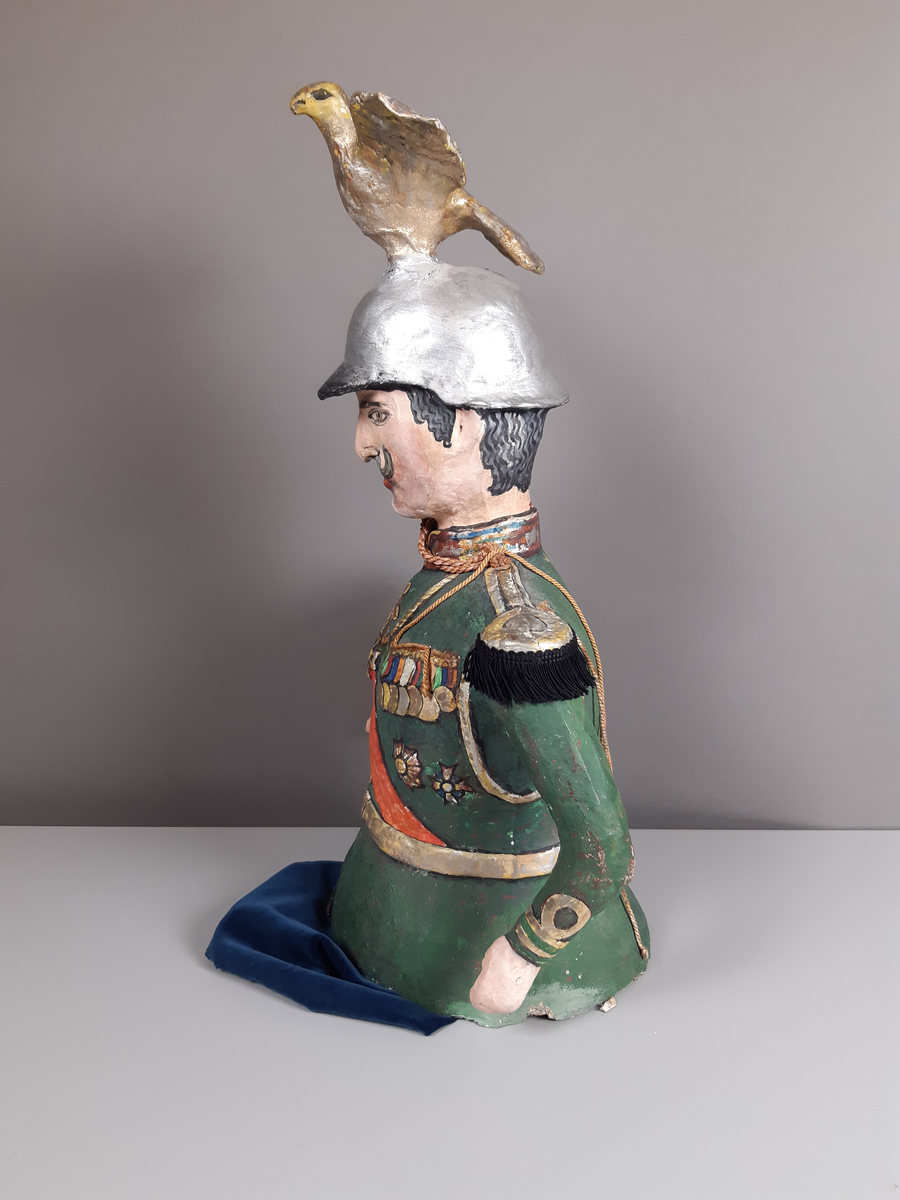 Docka i målad papier-maché, föreställande en man klädd i grön paraduniform och blank örnprydd hjälm. På bröstet har han en rad medaljer, ett rött ordensband över axeln, epåletter i guld med svarta tygfransar. Mannen har grått hår och mustasch. Byxorna består av mörkblå sammet.
