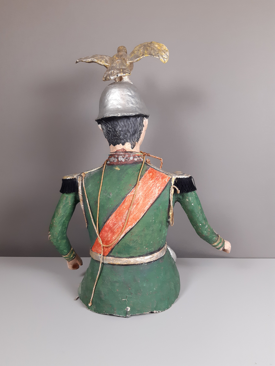 Docka i målad papier-maché, föreställande en man klädd i grön paraduniform och blank örnprydd hjälm. På bröstet har han en rad medaljer, ett rött ordensband över axeln, epåletter i guld med svarta tygfransar. Mannen har grått hår och mustasch. Byxorna består av mörkblå sammet.
