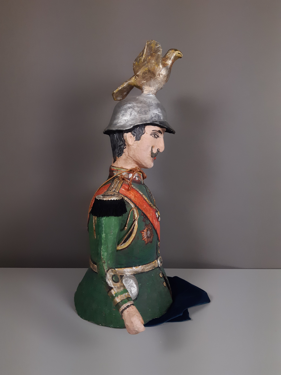 Docka i målad papier-maché, föreställande en man klädd i grön paraduniform och blank örnprydd hjälm. På bröstet har han en rad medaljer, ett rött ordensband över axeln, epåletter i guld med svarta tygfransar. Mannen har grått hår och mustasch. Byxorna består av mörkblå sammet.
