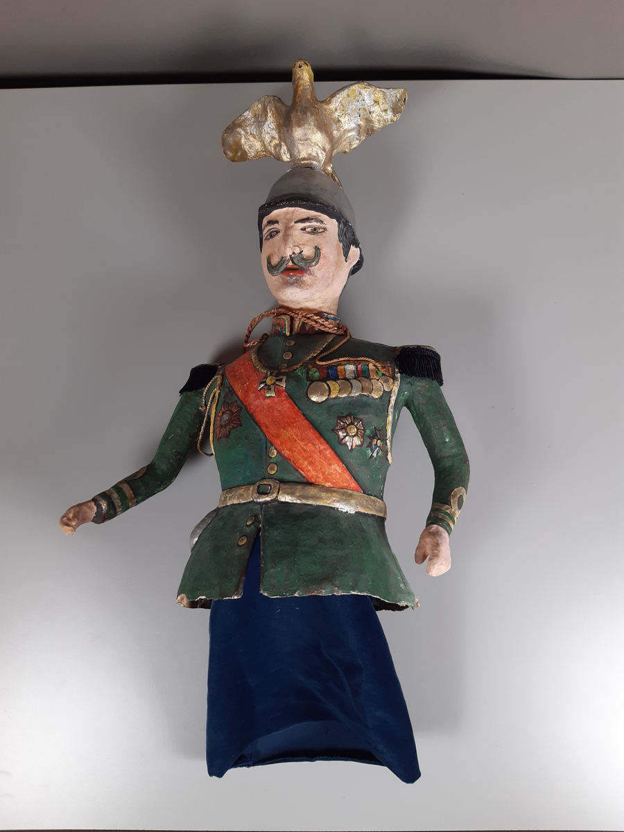 Docka i målad papier-maché, föreställande en man klädd i grön paraduniform och blank örnprydd hjälm. På bröstet har han en rad medaljer, ett rött ordensband över axeln, epåletter i guld med svarta tygfransar. Mannen har grått hår och mustasch. Byxorna består av mörkblå sammet.

