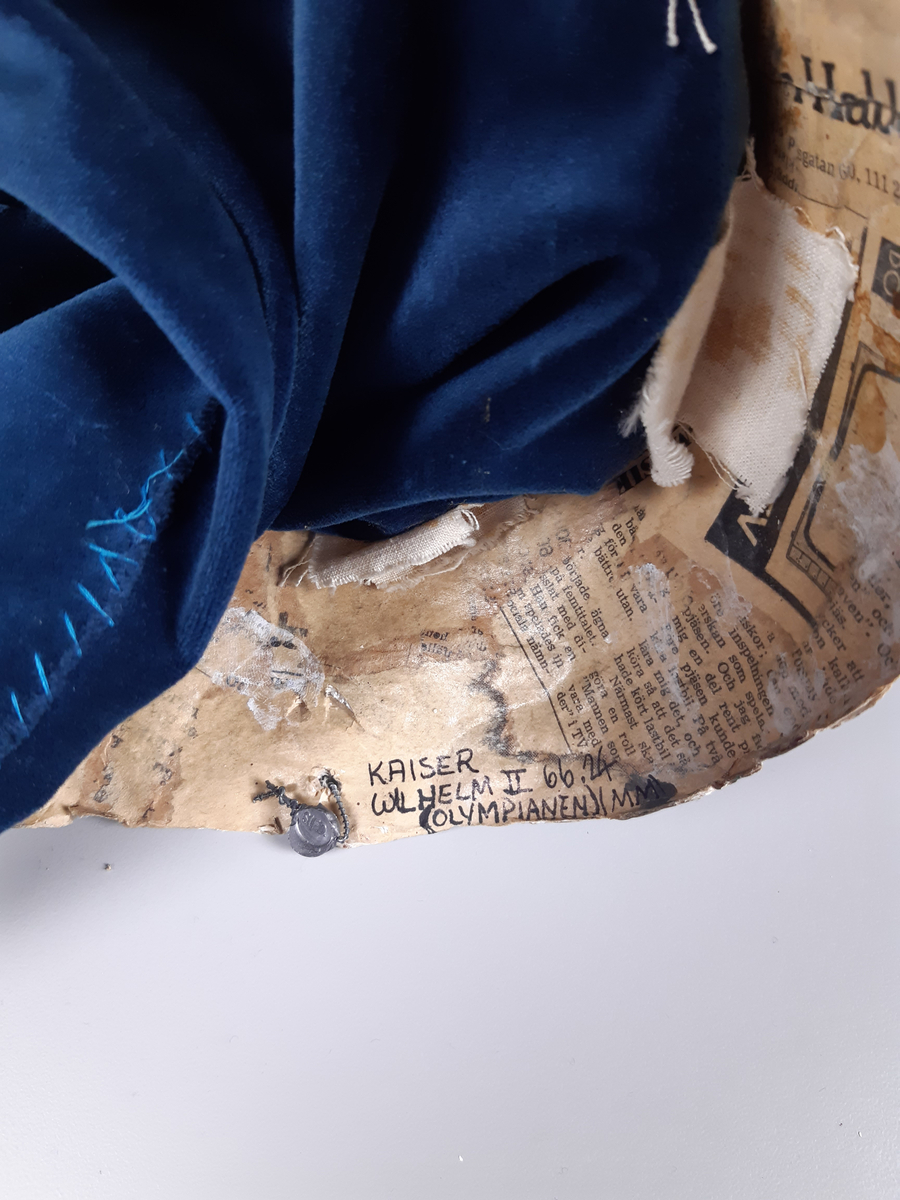 Docka i målad papier-maché, föreställande en man klädd i grön paraduniform och blank örnprydd hjälm. På bröstet har han en rad medaljer, ett rött ordensband över axeln, epåletter i guld med svarta tygfransar. Mannen har grått hår och mustasch. Byxorna består av mörkblå sammet.
