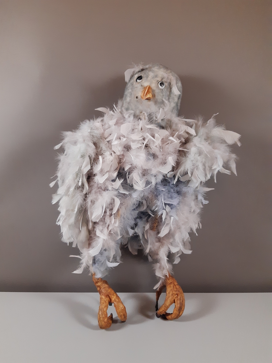 Stor grå uggla  med huvud av gips klädda i ljusgrå fjädrar och kropp av kraftig ståltråd klädd i en fjäderboa. Fötter och nädd är i gummiliknande material. Ugglan har ett plaströr i ryggen som handtag.
