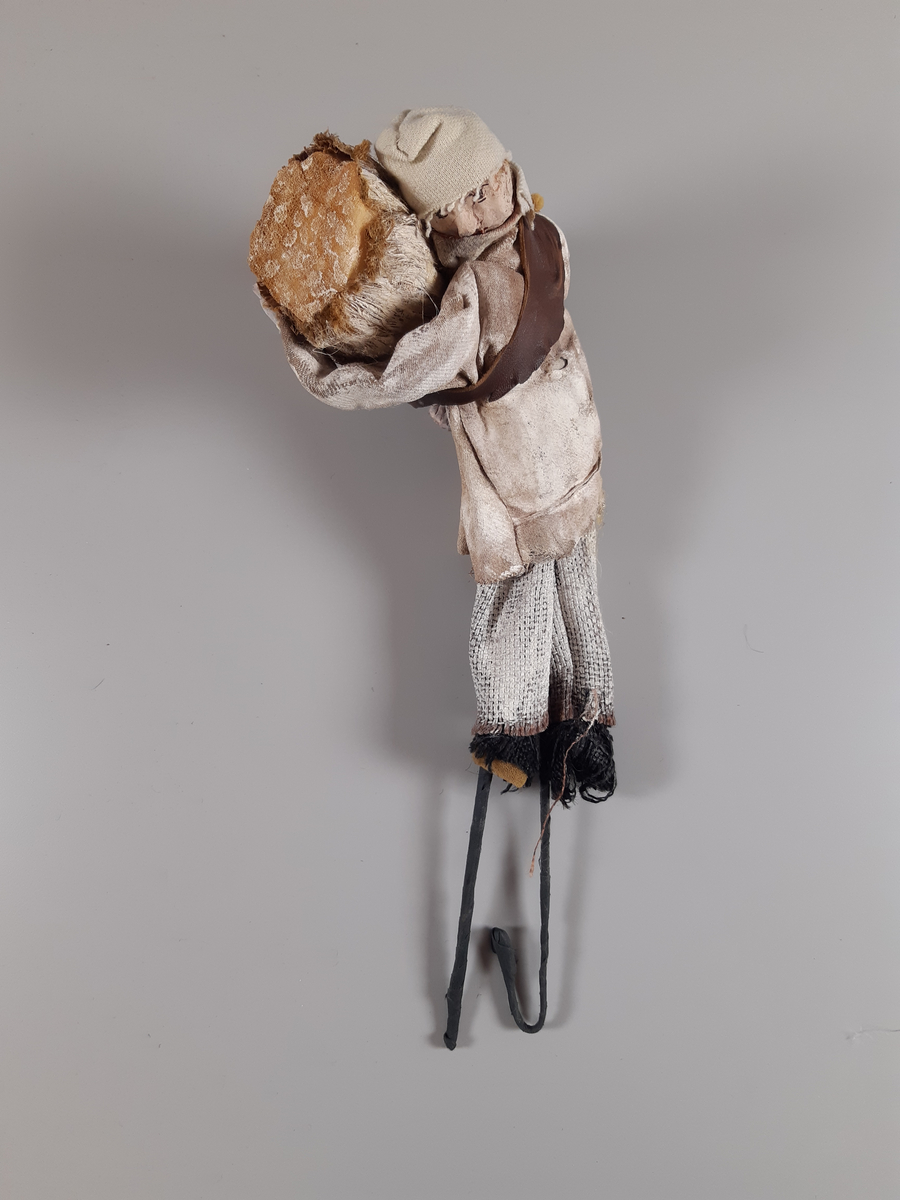 Stavdocka föreställande en man i vita kläder, långbyxor, skjorta och mössa, svarta skor och en brun bred läderrem över axel och rygg. Mannen bär vad som ser ut att vara ett stort fång ved i sina armar. Dockan manövreras med hjälp av två stavar i respektive fot.
