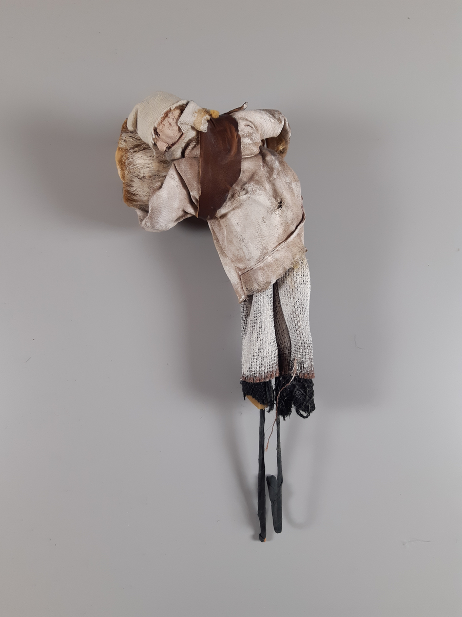 Stavdocka föreställande en man i vita kläder, långbyxor, skjorta och mössa, svarta skor och en brun bred läderrem över axel och rygg. Mannen bär vad som ser ut att vara ett stort fång ved i sina armar. Dockan manövreras med hjälp av två stavar i respektive fot.
