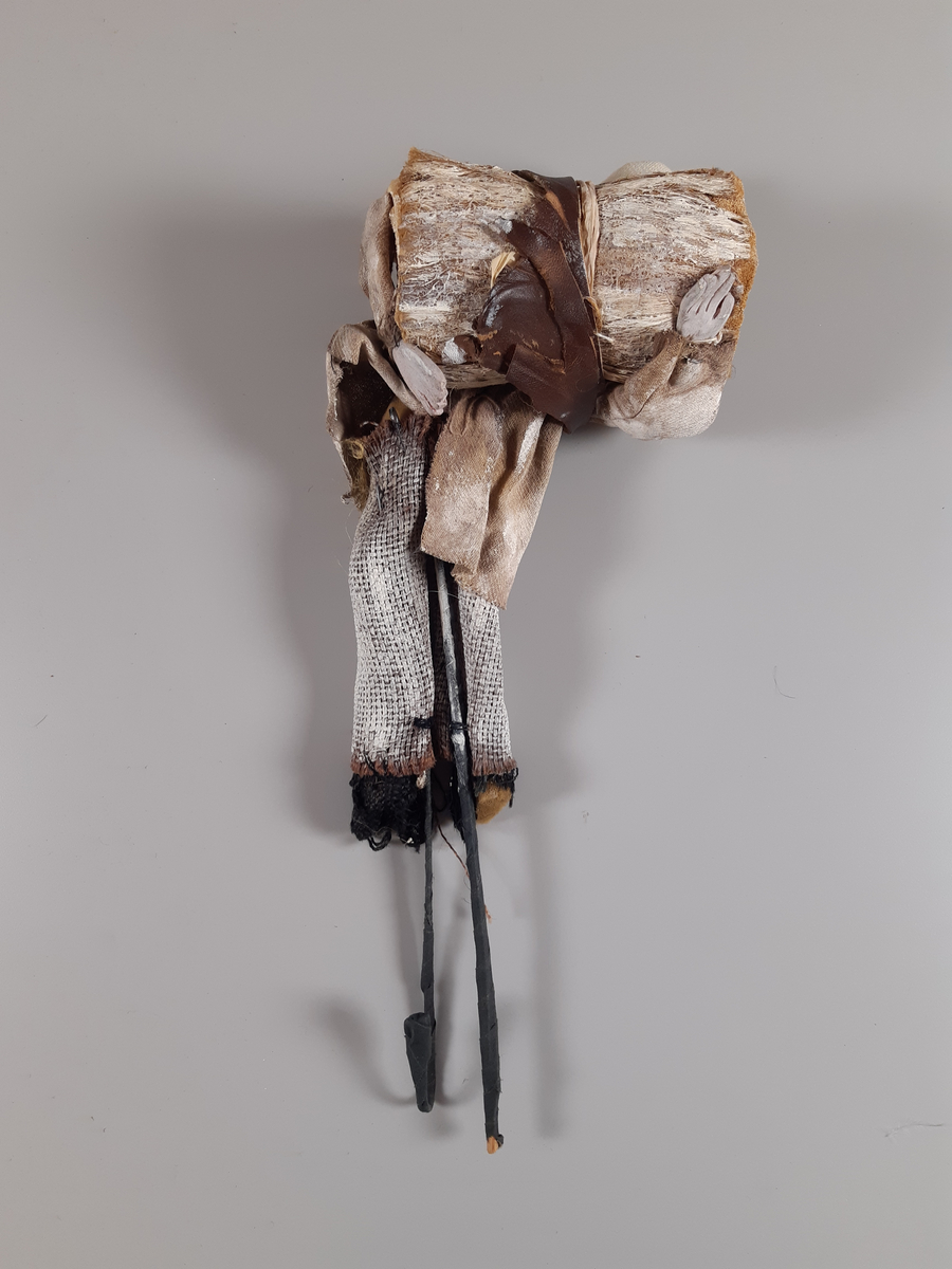 Stavdocka föreställande en man i vita kläder, långbyxor, skjorta och mössa, svarta skor och en brun bred läderrem över axel och rygg. Mannen bär vad som ser ut att vara ett stort fång ved i sina armar. Dockan manövreras med hjälp av två stavar i respektive fot.
