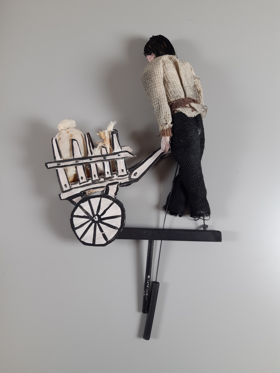 Stavdocka föreställande en man som dar en vagn. Han har mörkt hår och skägg, klädd i ljus skjorta, mörka byxor och har en gul skarf. Han håller i en vit vagn tillverkad av papp. På vagnen ligger två säckar av textil. Vagnen och mannen är fästade på en T-formad stav, det finns ytterligare en stav till en av mannens händer.
