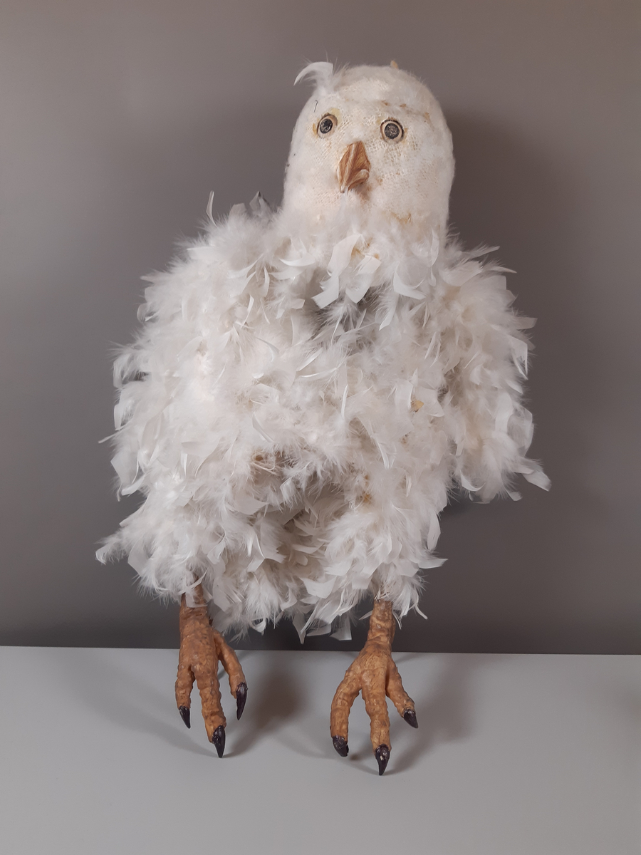 Stor vit uggla  med huvud av gips klädda i lvita fjädrar och kropp av kraftig ståltråd klädd i en fjäderboa. Fötter och nädd är i gummiliknande material. Ugglan har ett plaströr i ryggen som handtag.
