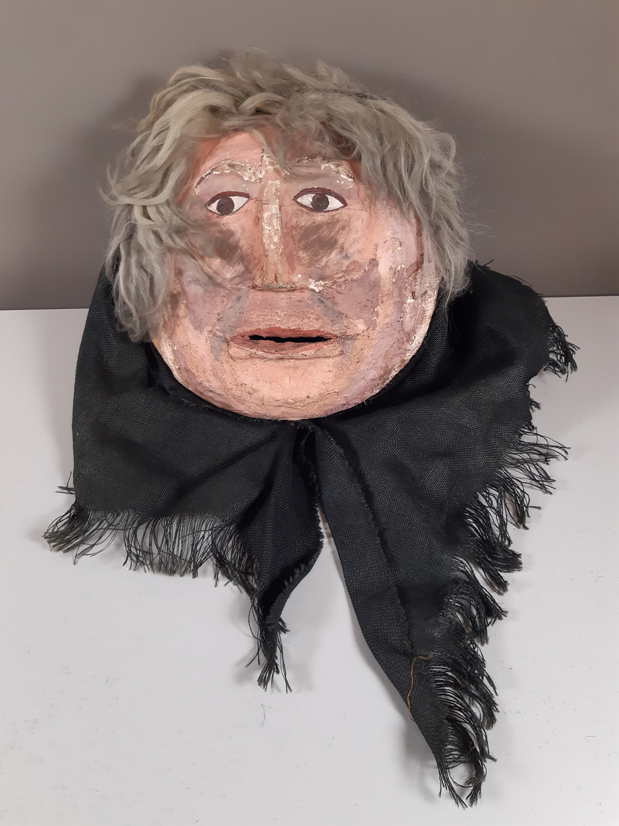 Mask föreställande äldre kvinna med svart huckle. Masken är monterad på en hockeyhjälm. 
