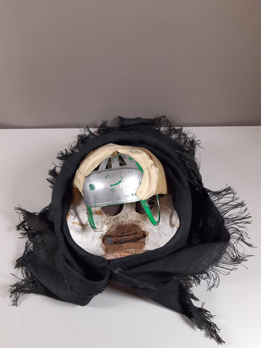 Mask föreställande äldre kvinna med svart huckle. Masken är monterad på en hockeyhjälm. 
