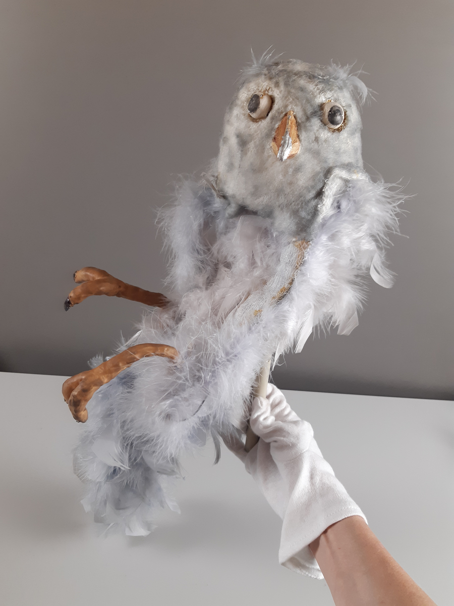 Uggla med huvud och vingar av gips, klädda i ljusgrå fjädrar, kroppen tillverkad av en fjäderboa. Fötter och nädd är i gummiliknande material. Ugglan har ett plaströr i ryggen som handtag samt en liten spak för att kunna röra ögonen från sida till sida.
