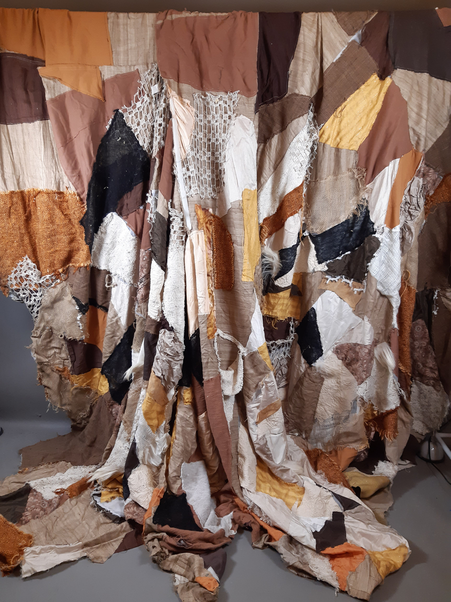 Stort tygstycke i lapptäcksteknik med tyglappar i rostbrunt, oramge och gult. "Klänningen" består av tre delar som går att fästa med kardborband.
