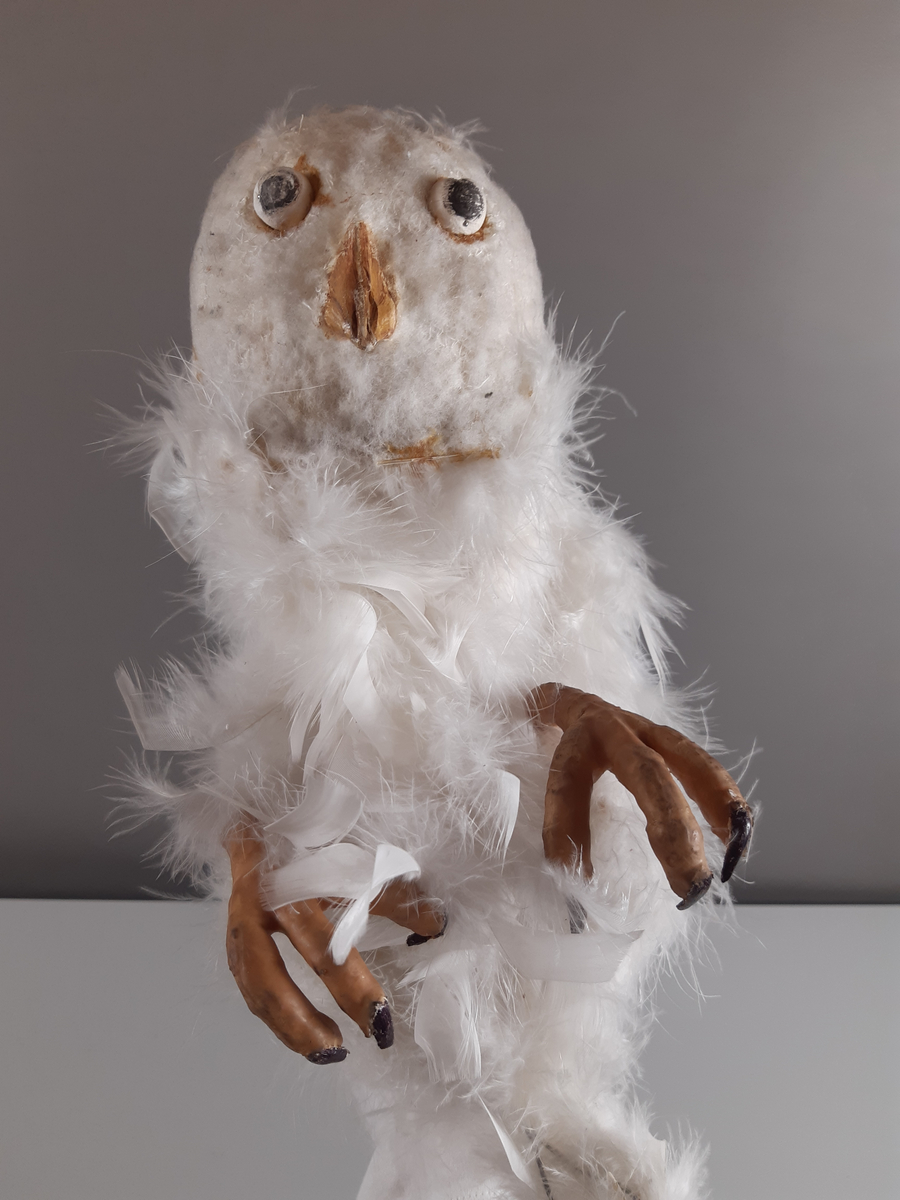 Uggla med huvud och vingar av gips, klädda i vita fjädrar, kroppen tillverkad av en fjäderboa. Fötter och nädd är i gummiliknande material. Ugglan har ett plaströr i ryggen som handtag samt en liten spak för att kunna röra ögonen från sida till sida.
