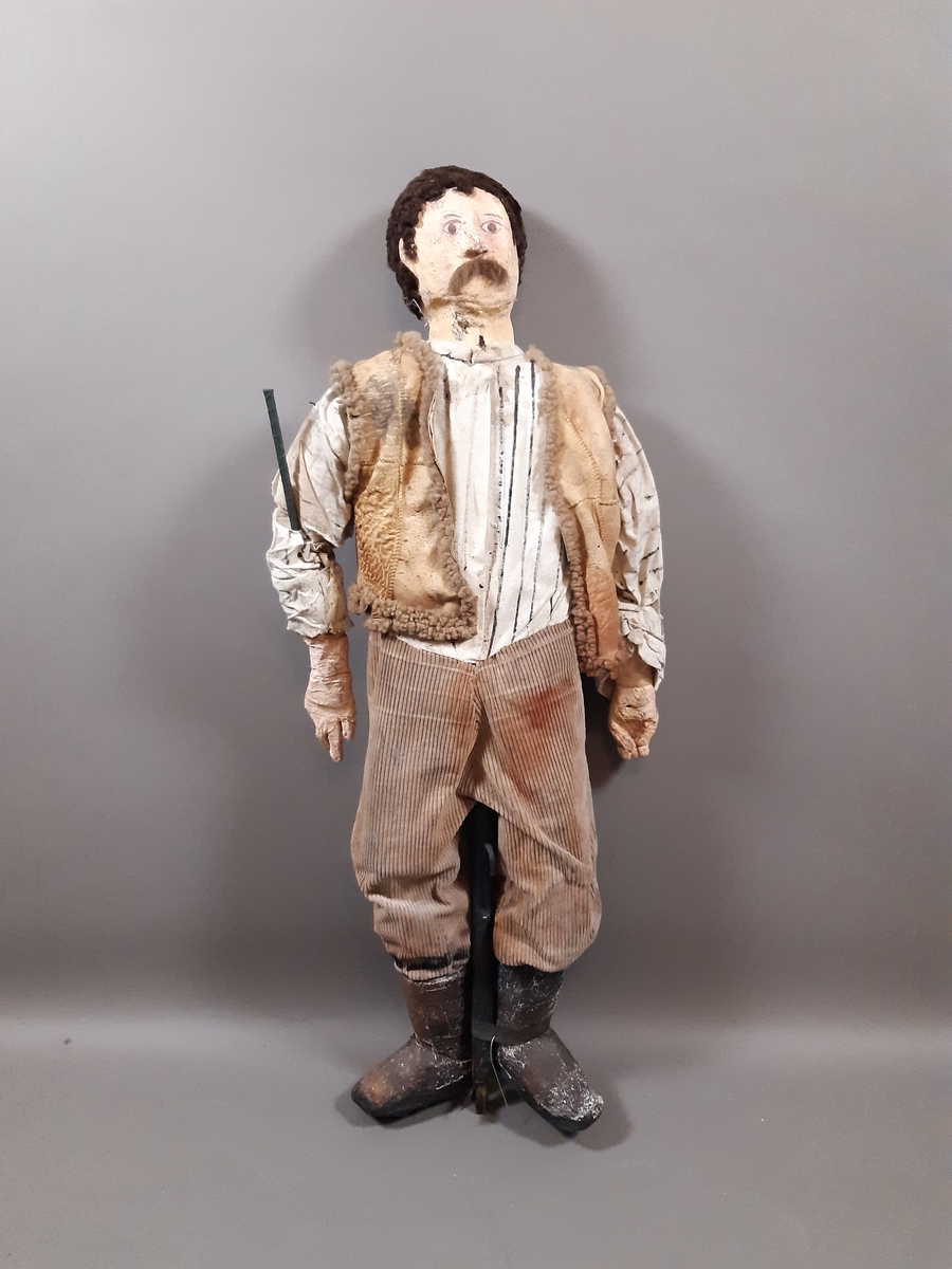 Speldocka föreställande en man med kort mörkt hår och mustasch klädd i höga stövlar, bruna långbyxor, vitrandig skjorta och ljusbrun väst. Dockan manövreras med hjälp av skaft i rygg, armar och fötter.

