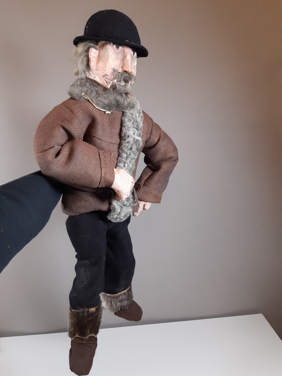 Speldocka föreställande en äldre man med grått hår och mustasch, klädd i mörka skinnstövlar, mörka långbyxor, mörkbrun tjock jacka med grå päls och svart plommonstop. Dockan manövreras med hjälp av skaft i rygg, armar och fötter.
