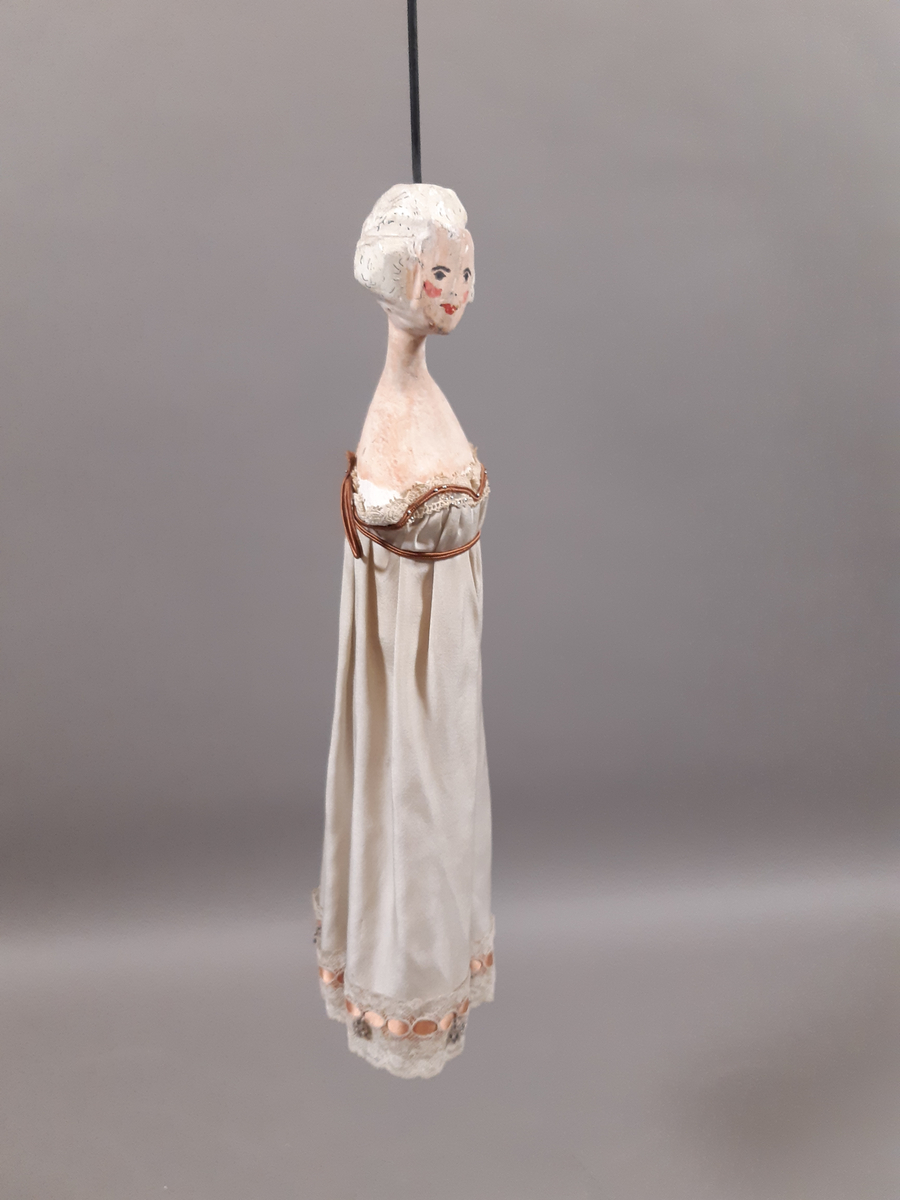 Föreställer en kvinna med vitt hår klädd i vit empirklänning. Dockans armar är lösa. Marionett går att fästa ihop med den andra dockan i dansparet med hjälp av en metallplatta.
