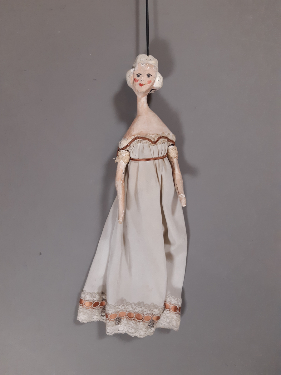 Föreställer en kvinna med vitt hår klädd i vit empirklänning. Dockans armar är lösa. Marionett går att fästa ihop med den andra dockan i dansparet med hjälp av en metallplatta.
