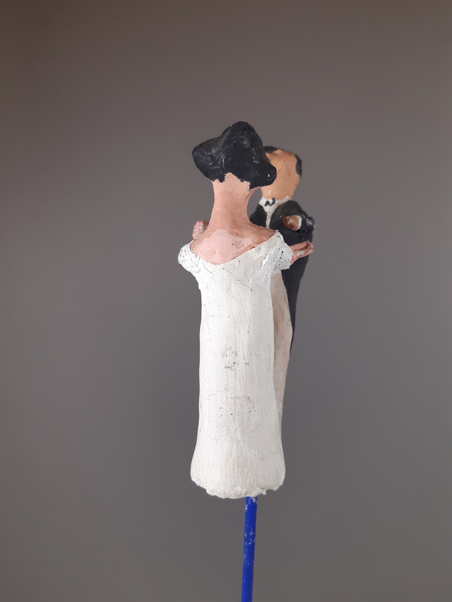 Föreställer en kvinna klädd i vit empirklänning och en man klädd i svart frackjacka, höghalsad vit skjorta och ljusa långbyxor. Både mannen och kvinnan har mörkt hår.
