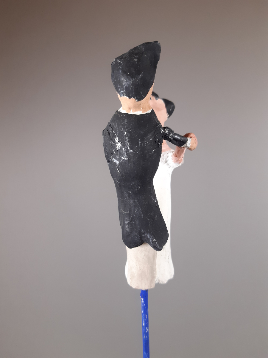 Föreställer en kvinna klädd i vit empirklänning och en man klädd i svart frackjacka, höghalsad vit skjorta och ljusa långbyxor. Både mannen och kvinnan har mörkt hår.
