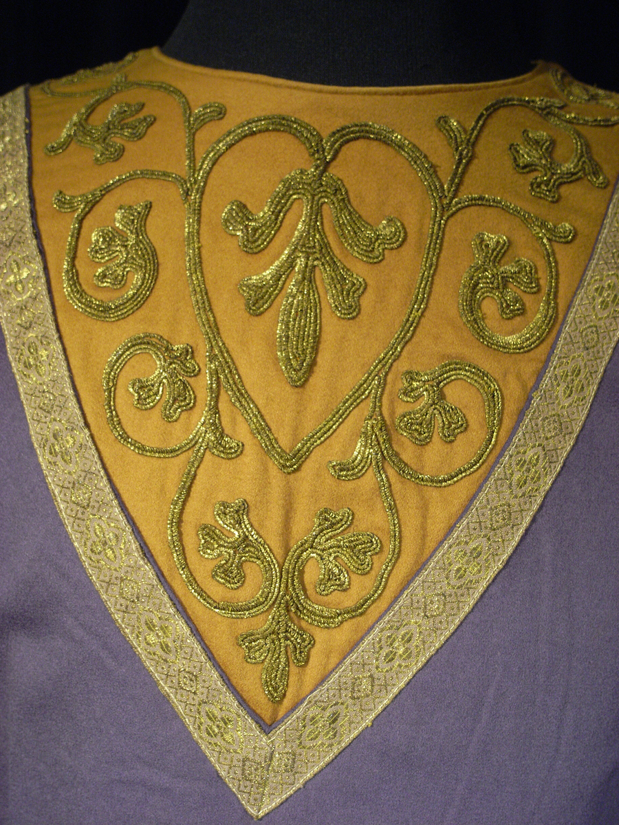 Tunika av fint ylletyg i mörklila färg. Tunikan är något längre med djupa sprund i vardera sida och långa vida ärmar med underliggande kroppsnära ärmar av fint ylletyg i senapsgul färg. Dekor runt halsringning som avslutas framtill i ett V av ylletyg i senapsgul färg dekorerad med stiliserade växtslingor av metalltråd i guldfärg. Applikationsdekor i form av växtslingor längs nederkanten av ylletyg i mörkgrå färg kantat med metalltråd i guldfärg. Ärmsluten på de underliggande ärmarna liknande dekorerade med applikation av ylletyg i brun färg kantat med metalltråd i guldfärg. I höger sidosprund en snodd i guldfärg som delvis lossnat. Längre sprund mitt bak med dold knäppning med runda metallknappar som lagts ut med ett sammetstyg i mörklila färg. Fodrad med linnetyg i vit färg.
Saknas snodd i vänster sidosprund. Snodd i höger sprund har delvis lossnat. Flertal knappar saknas.