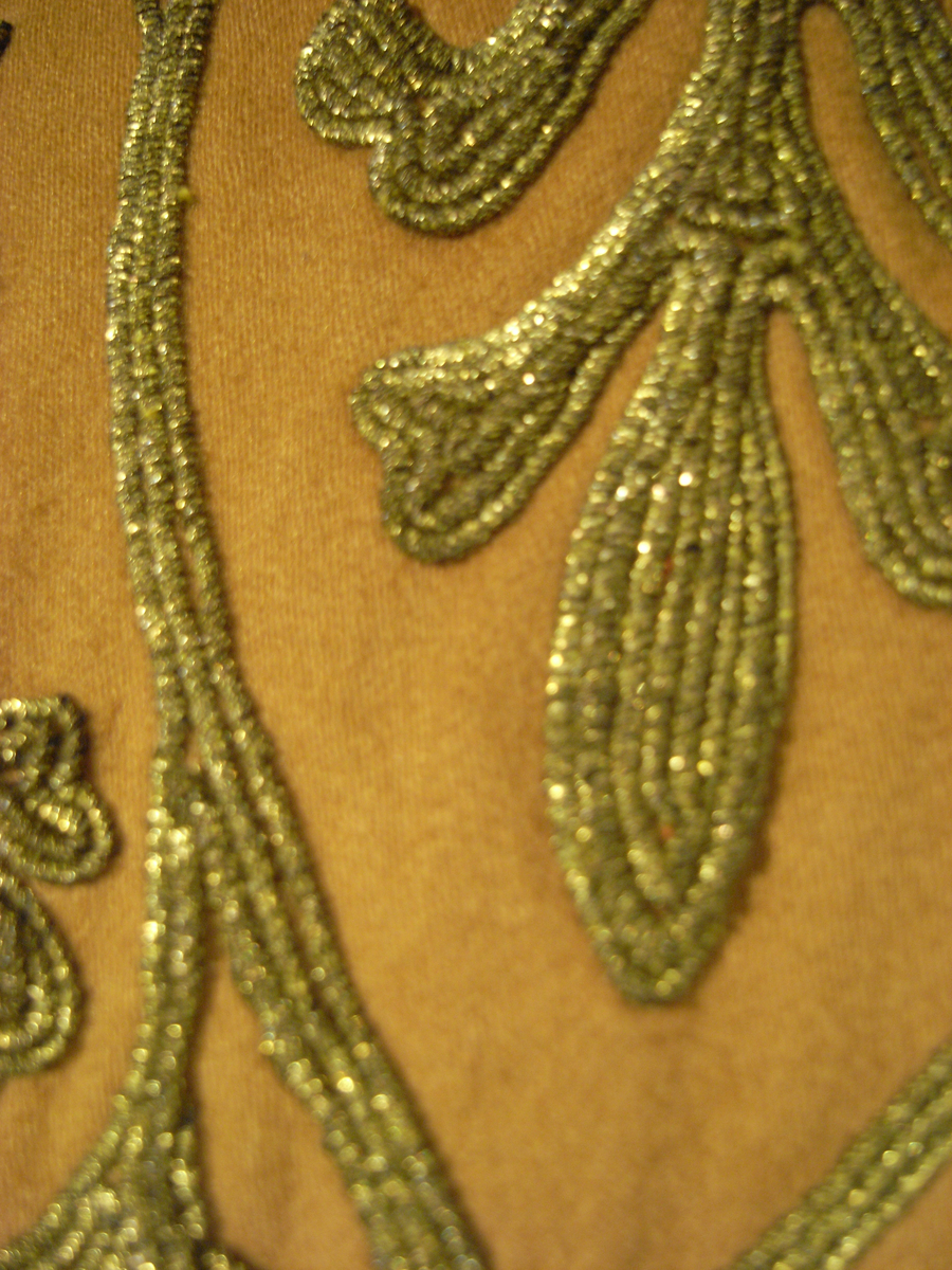 Tunika av fint ylletyg i mörklila färg. Tunikan är något längre med djupa sprund i vardera sida och långa vida ärmar med underliggande kroppsnära ärmar av fint ylletyg i senapsgul färg. Dekor runt halsringning som avslutas framtill i ett V av ylletyg i senapsgul färg dekorerad med stiliserade växtslingor av metalltråd i guldfärg. Applikationsdekor i form av växtslingor längs nederkanten av ylletyg i mörkgrå färg kantat med metalltråd i guldfärg. Ärmsluten på de underliggande ärmarna liknande dekorerade med applikation av ylletyg i brun färg kantat med metalltråd i guldfärg. I höger sidosprund en snodd i guldfärg som delvis lossnat. Längre sprund mitt bak med dold knäppning med runda metallknappar som lagts ut med ett sammetstyg i mörklila färg. Fodrad med linnetyg i vit färg.
Saknas snodd i vänster sidosprund. Snodd i höger sprund har delvis lossnat. Flertal knappar saknas.