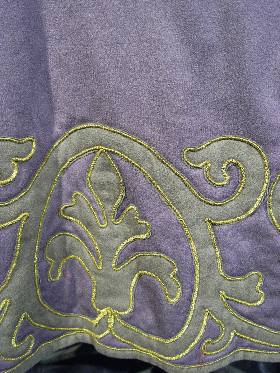 Tunika av fint ylletyg i mörklila färg. Tunikan är något längre med djupa sprund i vardera sida och långa vida ärmar med underliggande kroppsnära ärmar av fint ylletyg i senapsgul färg. Dekor runt halsringning som avslutas framtill i ett V av ylletyg i senapsgul färg dekorerad med stiliserade växtslingor av metalltråd i guldfärg. Applikationsdekor i form av växtslingor längs nederkanten av ylletyg i mörkgrå färg kantat med metalltråd i guldfärg. Ärmsluten på de underliggande ärmarna liknande dekorerade med applikation av ylletyg i brun färg kantat med metalltråd i guldfärg. I höger sidosprund en snodd i guldfärg som delvis lossnat. Längre sprund mitt bak med dold knäppning med runda metallknappar som lagts ut med ett sammetstyg i mörklila färg. Fodrad med linnetyg i vit färg.
Saknas snodd i vänster sidosprund. Snodd i höger sprund har delvis lossnat. Flertal knappar saknas.