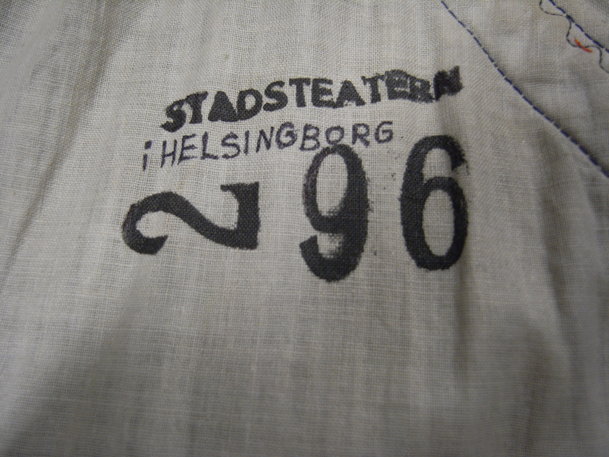 Tunika av fint ylletyg i mörklila färg. Tunikan är något längre med djupa sprund i vardera sida och långa vida ärmar med underliggande kroppsnära ärmar av fint ylletyg i senapsgul färg. Dekor runt halsringning som avslutas framtill i ett V av ylletyg i senapsgul färg dekorerad med stiliserade växtslingor av metalltråd i guldfärg. Applikationsdekor i form av växtslingor längs nederkanten av ylletyg i mörkgrå färg kantat med metalltråd i guldfärg. Ärmsluten på de underliggande ärmarna liknande dekorerade med applikation av ylletyg i brun färg kantat med metalltråd i guldfärg. I höger sidosprund en snodd i guldfärg som delvis lossnat. Längre sprund mitt bak med dold knäppning med runda metallknappar som lagts ut med ett sammetstyg i mörklila färg. Fodrad med linnetyg i vit färg.
Saknas snodd i vänster sidosprund. Snodd i höger sprund har delvis lossnat. Flertal knappar saknas.