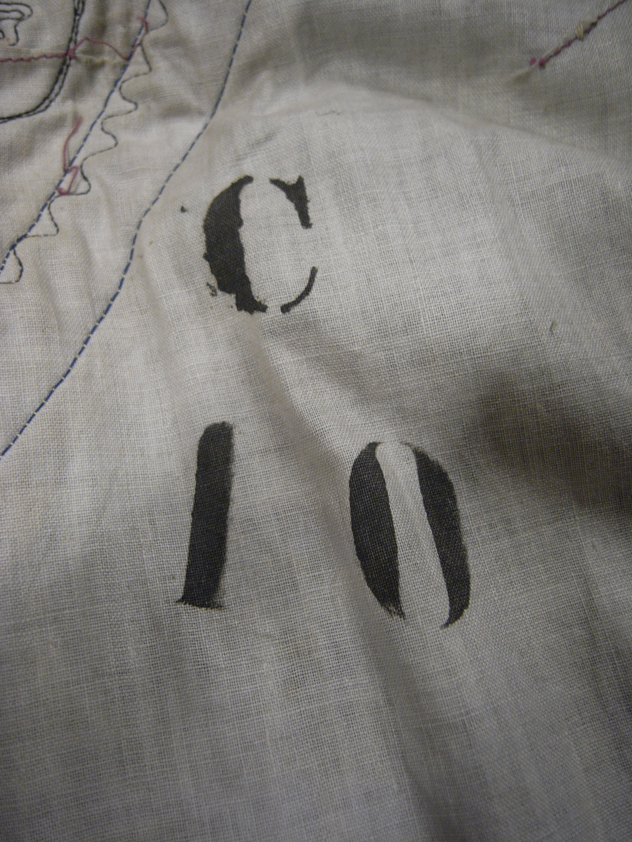 Tunika av fint ylletyg i mörklila färg. Tunikan är något längre med djupa sprund i vardera sida och långa vida ärmar med underliggande kroppsnära ärmar av fint ylletyg i senapsgul färg. Dekor runt halsringning som avslutas framtill i ett V av ylletyg i senapsgul färg dekorerad med stiliserade växtslingor av metalltråd i guldfärg. Applikationsdekor i form av växtslingor längs nederkanten av ylletyg i mörkgrå färg kantat med metalltråd i guldfärg. Ärmsluten på de underliggande ärmarna liknande dekorerade med applikation av ylletyg i brun färg kantat med metalltråd i guldfärg. I höger sidosprund en snodd i guldfärg som delvis lossnat. Längre sprund mitt bak med dold knäppning med runda metallknappar som lagts ut med ett sammetstyg i mörklila färg. Fodrad med linnetyg i vit färg.
Saknas snodd i vänster sidosprund. Snodd i höger sprund har delvis lossnat. Flertal knappar saknas.