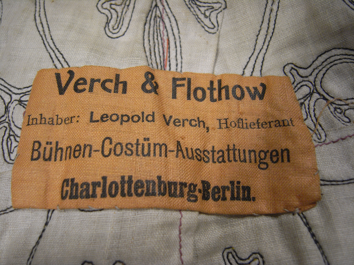 Tunika av fint ylletyg i mörklila färg. Tunikan är något längre med djupa sprund i vardera sida och långa vida ärmar med underliggande kroppsnära ärmar av fint ylletyg i senapsgul färg. Dekor runt halsringning som avslutas framtill i ett V av ylletyg i senapsgul färg dekorerad med stiliserade växtslingor av metalltråd i guldfärg. Applikationsdekor i form av växtslingor längs nederkanten av ylletyg i mörkgrå färg kantat med metalltråd i guldfärg. Ärmsluten på de underliggande ärmarna liknande dekorerade med applikation av ylletyg i brun färg kantat med metalltråd i guldfärg. I höger sidosprund en snodd i guldfärg som delvis lossnat. Längre sprund mitt bak med dold knäppning med runda metallknappar som lagts ut med ett sammetstyg i mörklila färg. Fodrad med linnetyg i vit färg.
Saknas snodd i vänster sidosprund. Snodd i höger sprund har delvis lossnat. Flertal knappar saknas.