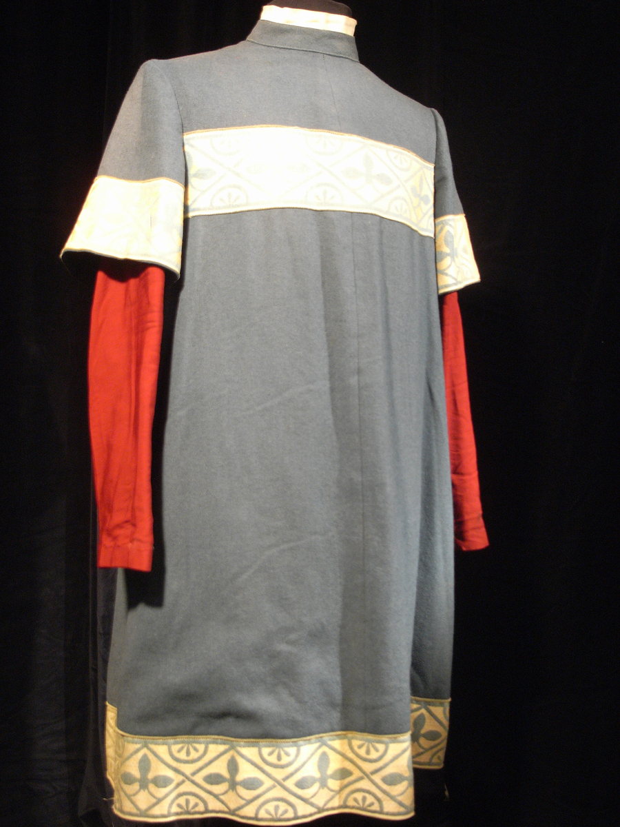 Tunika/kolt av ulltyg, kläde, i duvblå fräg. En bred bård av ett mönstervävt tyg i vit och ljusblå färg längs nederkant, ärmar och bål. Långa ärmar av ett tyg i röd färg. 

