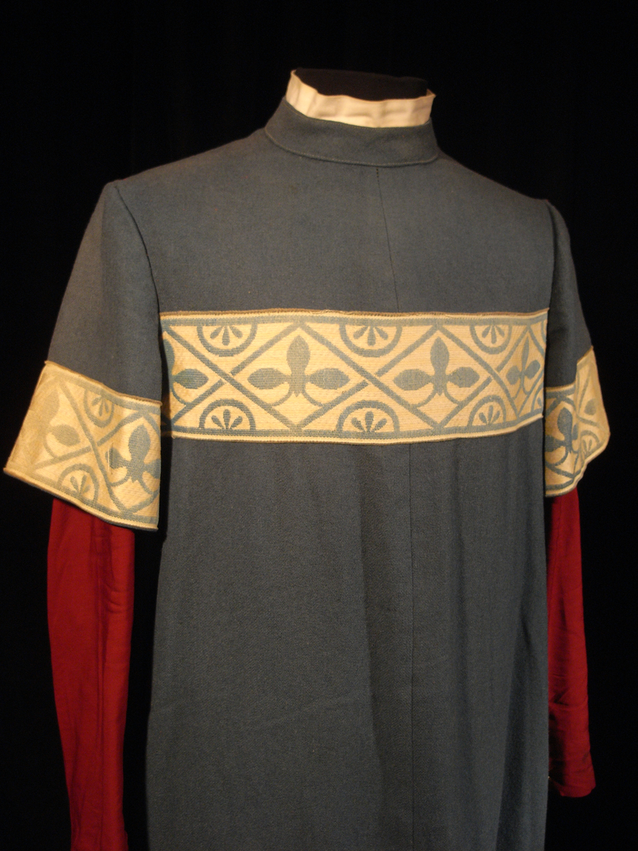 Tunika/kolt av ulltyg, kläde, i duvblå fräg. En bred bård av ett mönstervävt tyg i vit och ljusblå färg längs nederkant, ärmar och bål. Långa ärmar av ett tyg i röd färg. 

