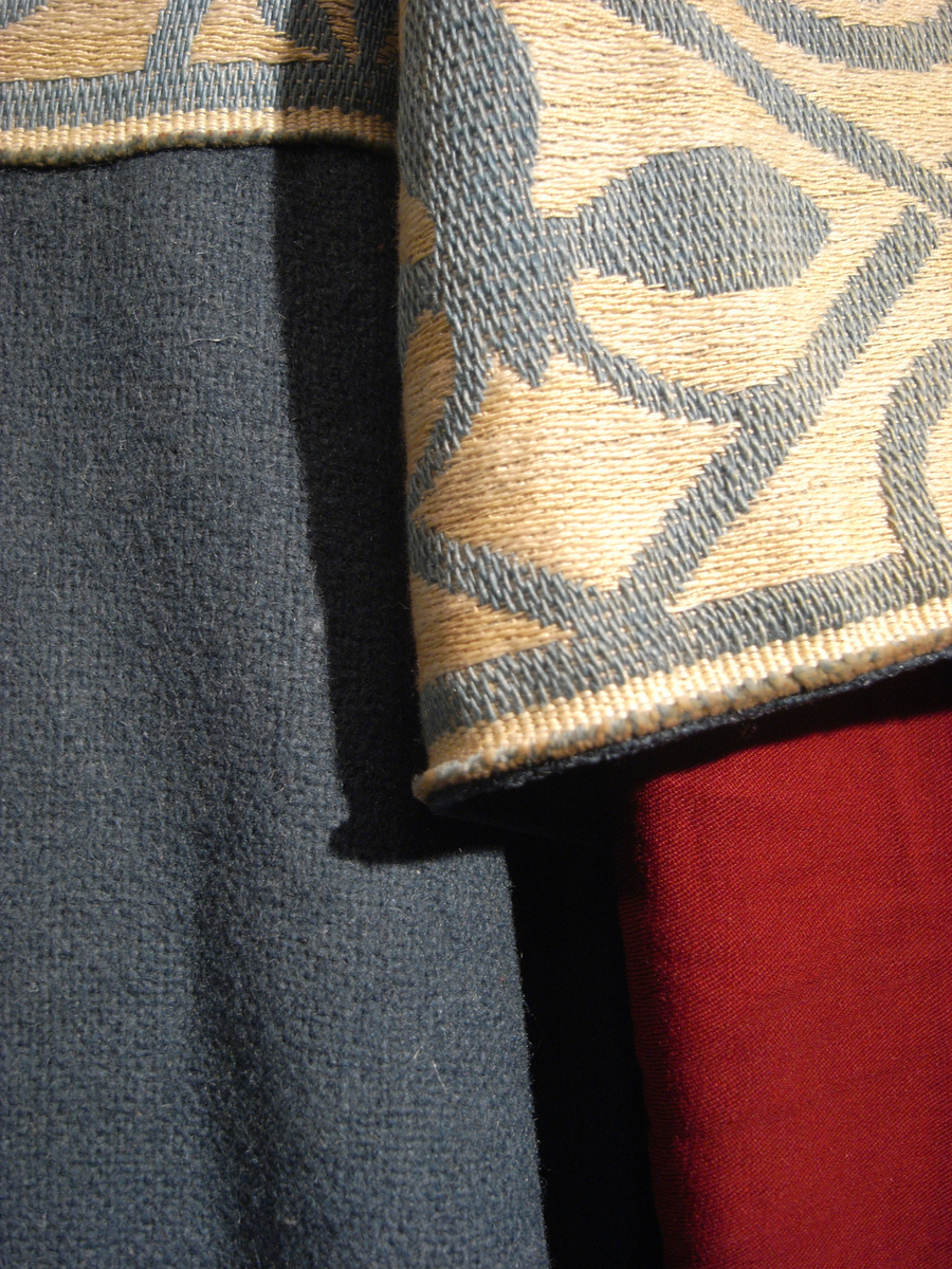 Tunika/kolt av ulltyg, kläde, i duvblå fräg. En bred bård av ett mönstervävt tyg i vit och ljusblå färg längs nederkant, ärmar och bål. Långa ärmar av ett tyg i röd färg. 
