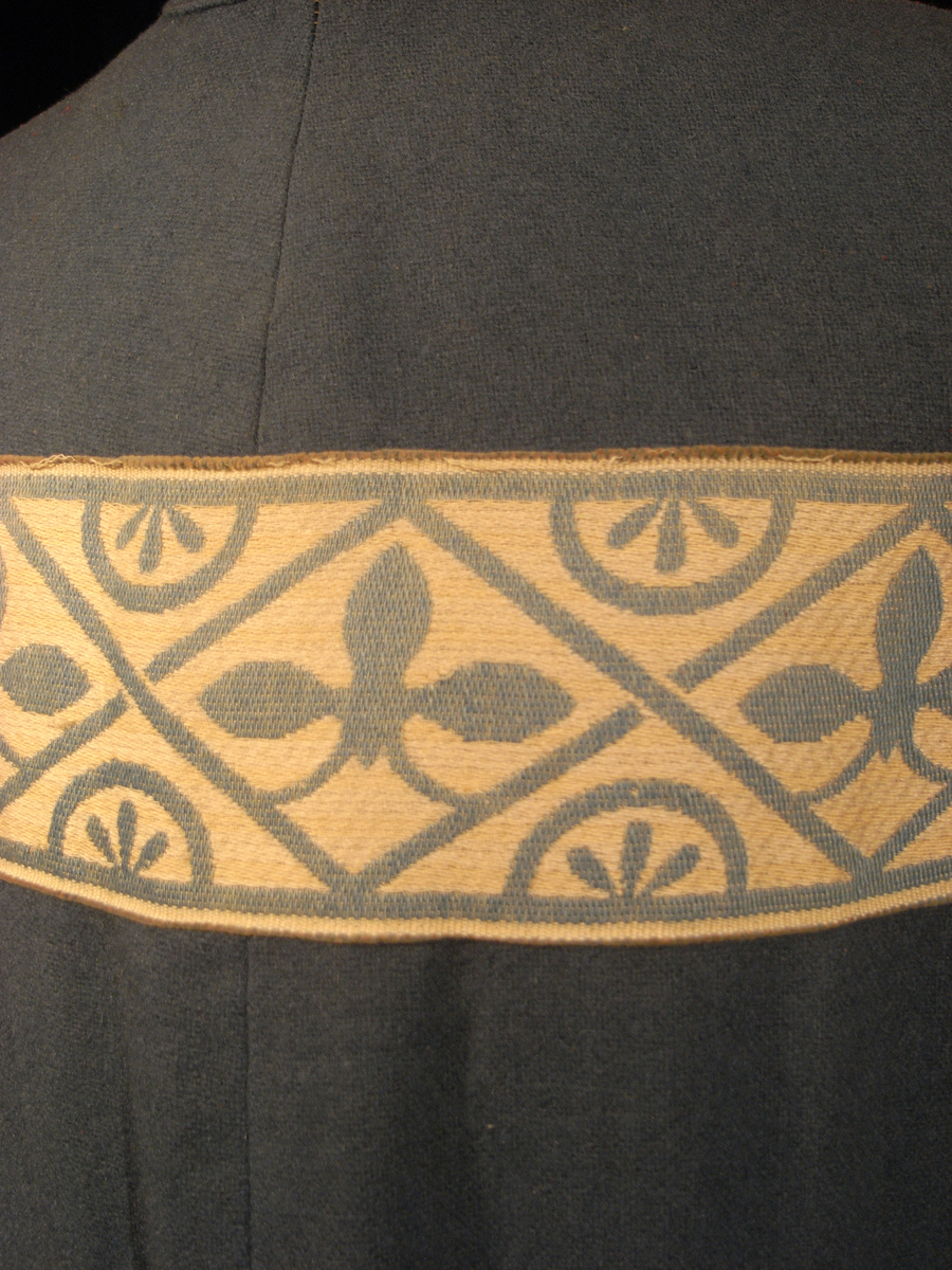 Tunika/kolt av ulltyg, kläde, i duvblå fräg. En bred bård av ett mönstervävt tyg i vit och ljusblå färg längs nederkant, ärmar och bål. Långa ärmar av ett tyg i röd färg. 
