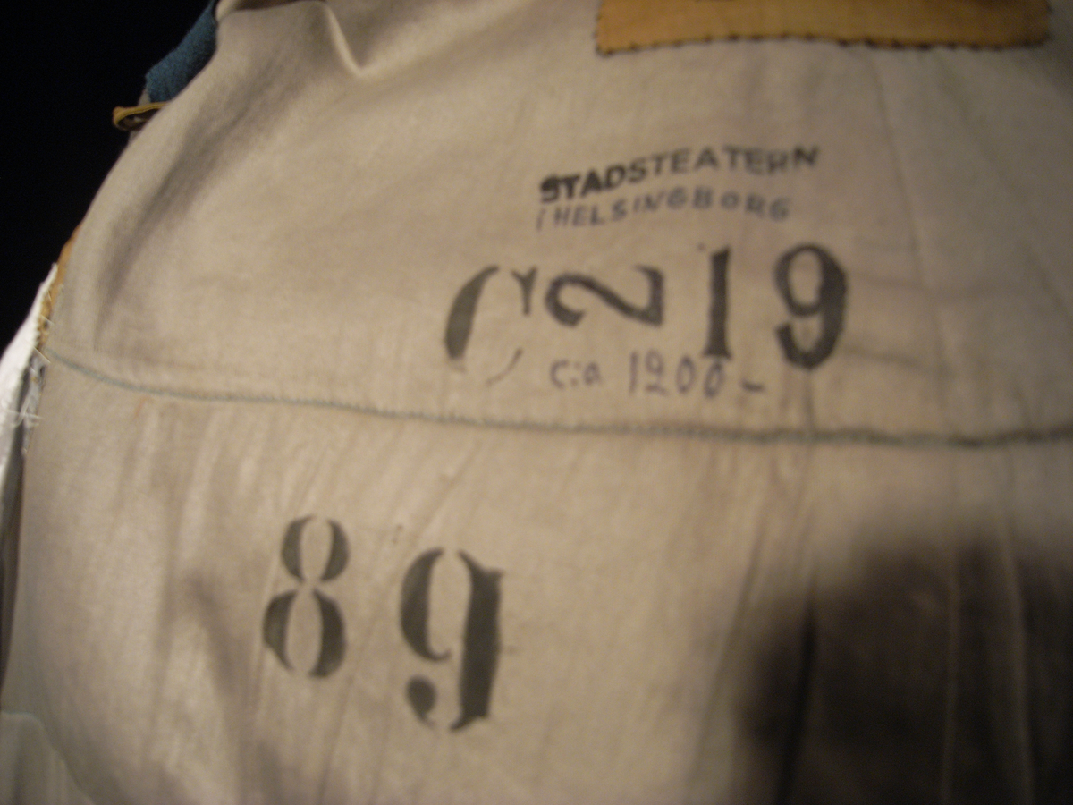 Tunika/kolt av ulltyg, kläde, i duvblå fräg. En bred bård av ett mönstervävt tyg i vit och ljusblå färg längs nederkant, ärmar och bål. Långa ärmar av ett tyg i röd färg. 
