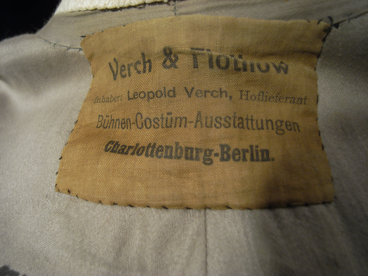 Tunika/kolt av ulltyg, kläde, i duvblå fräg. En bred bård av ett mönstervävt tyg i vit och ljusblå färg längs nederkant, ärmar och bål. Långa ärmar av ett tyg i röd färg. 
